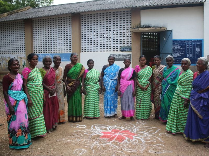 A group of twelve older women standing side-by-side in a semi-circle outdoors in Tamil Nadu, India. They are dressed in bright, colorful traditional saris in shades of pink, green, blue, purple, and yellow with various striped, patterned, and solid designs. In front of them on the dirt ground is a large geometric rangoli pattern drawn in white and red chalk or powder. Behind the group is a building featuring white lattice walls, a blue door, and signs written in Tamil script on the wall.